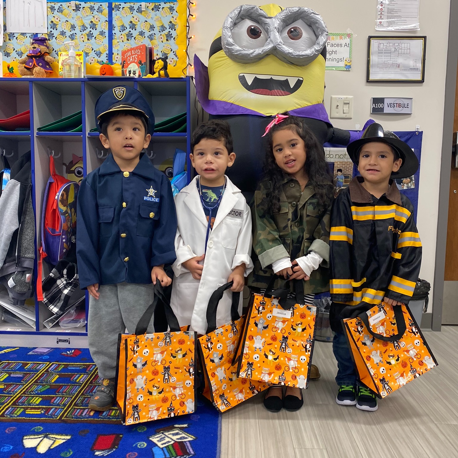 Four children in costumes stand in front of a classroom cubby area, each holding an orange Halloween-themed tote bag.