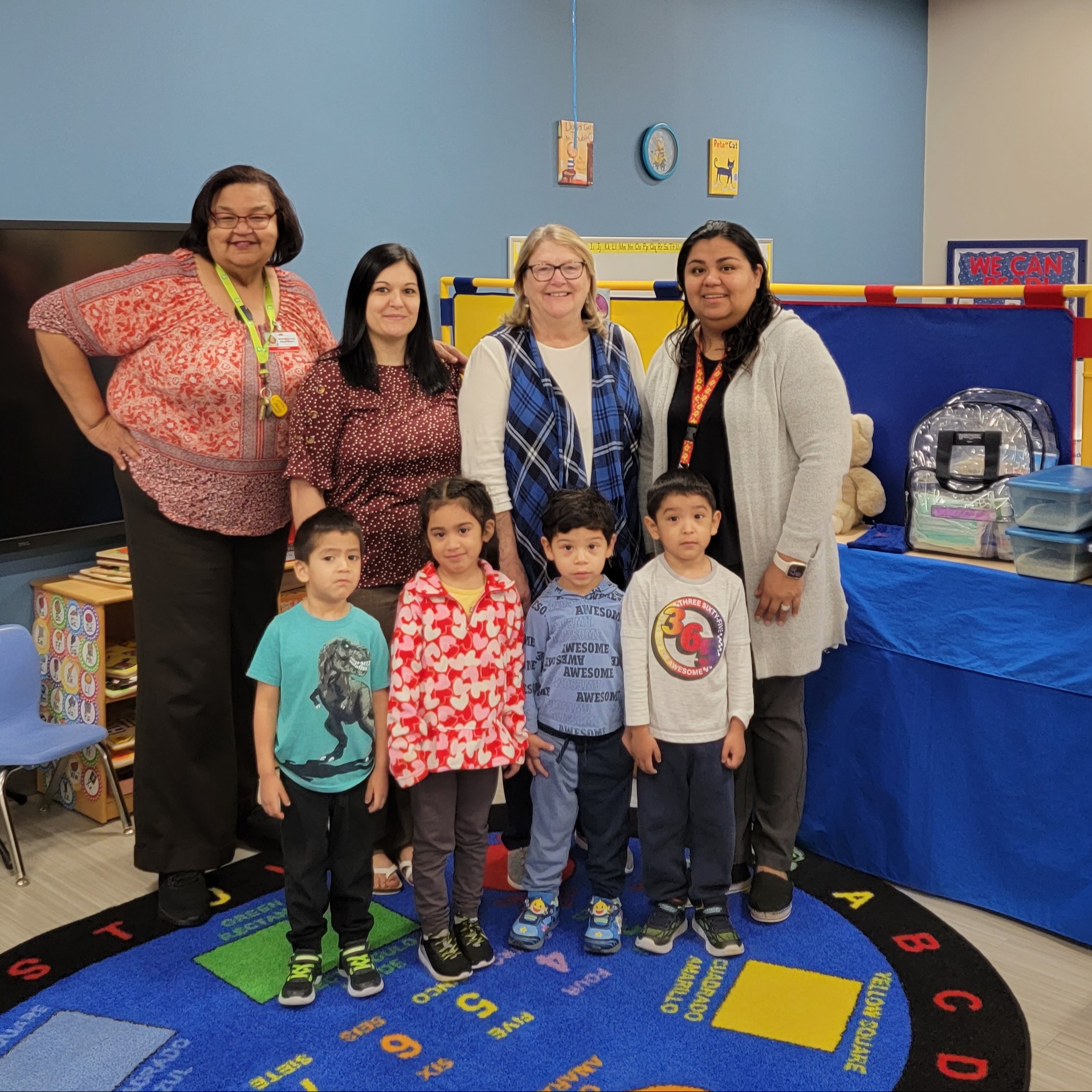 Group photo in a classroom with five children standing in front of four adults, surrounded by educational items and decorations.