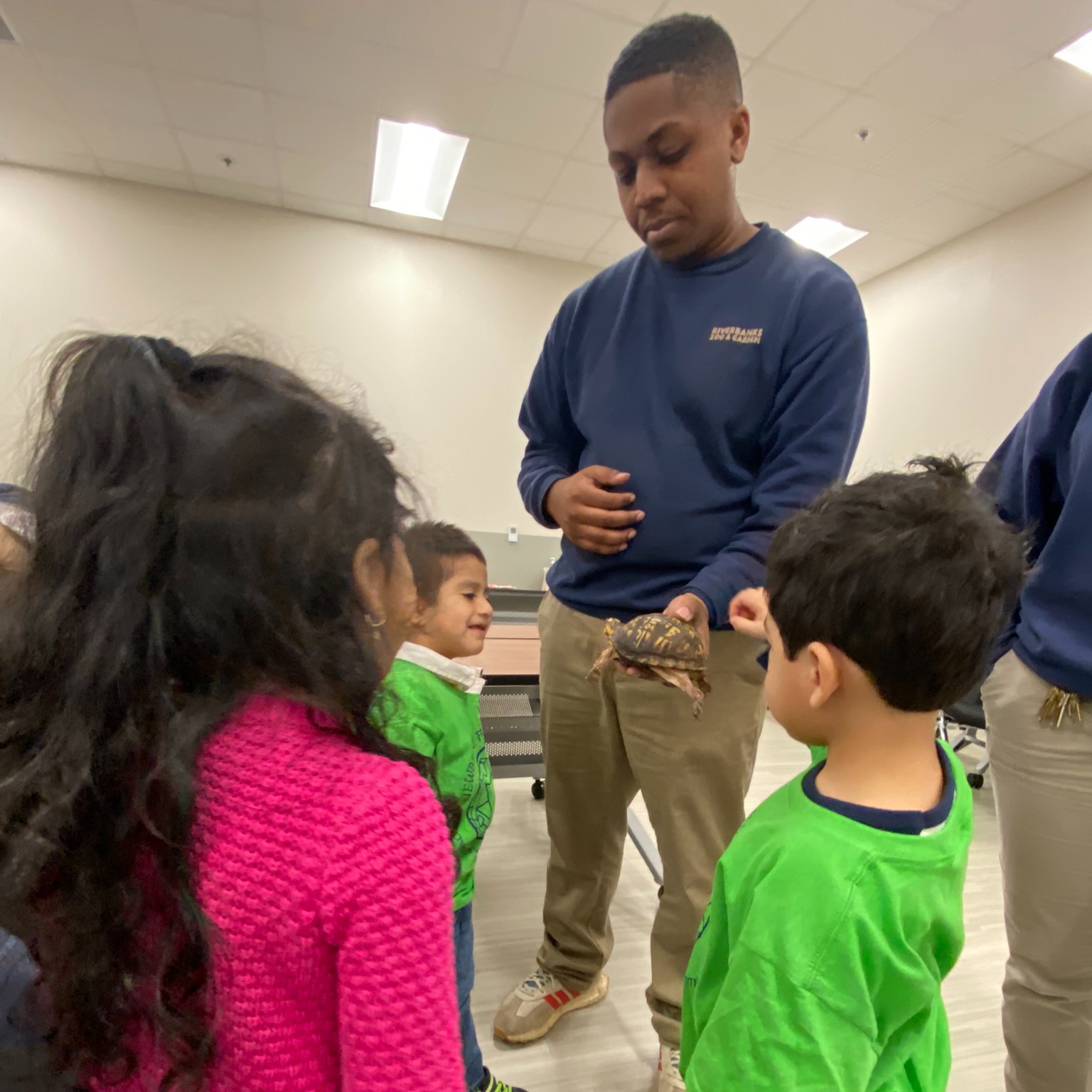 An adult holding a turtle shows it to three young children in a classroom setting.