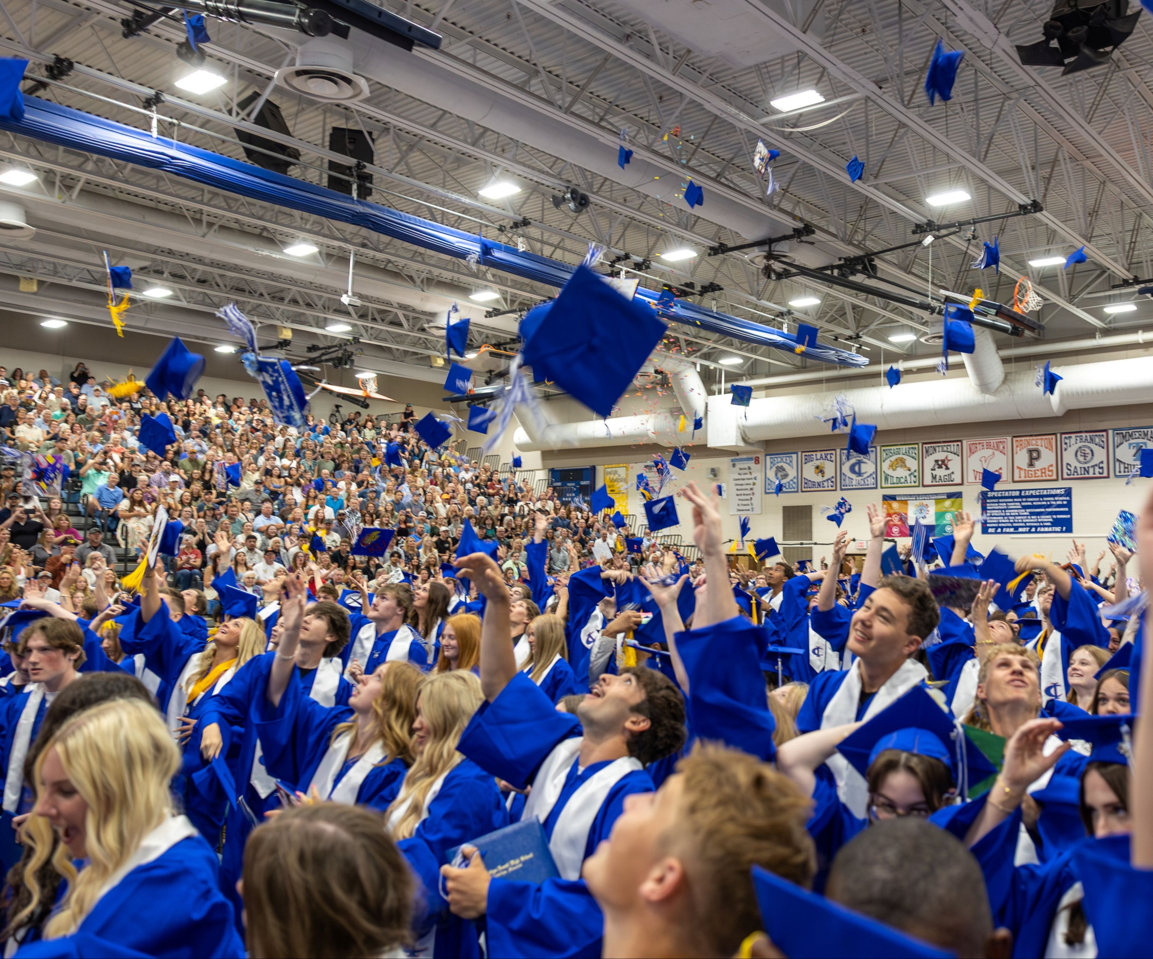 Graduates in blue gowns celebrate by throwing their caps into the air during a graduation ceremony in a gymnasium.