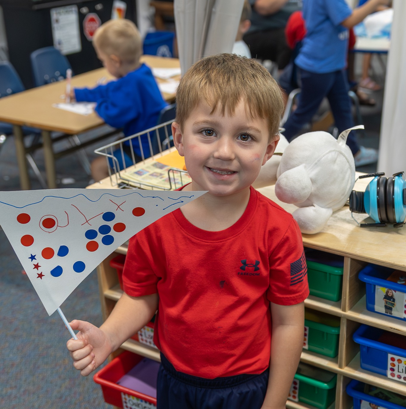 A smiling young student in a red shirt stands in a classroom holding up a hand-drawn paper pennant flag.