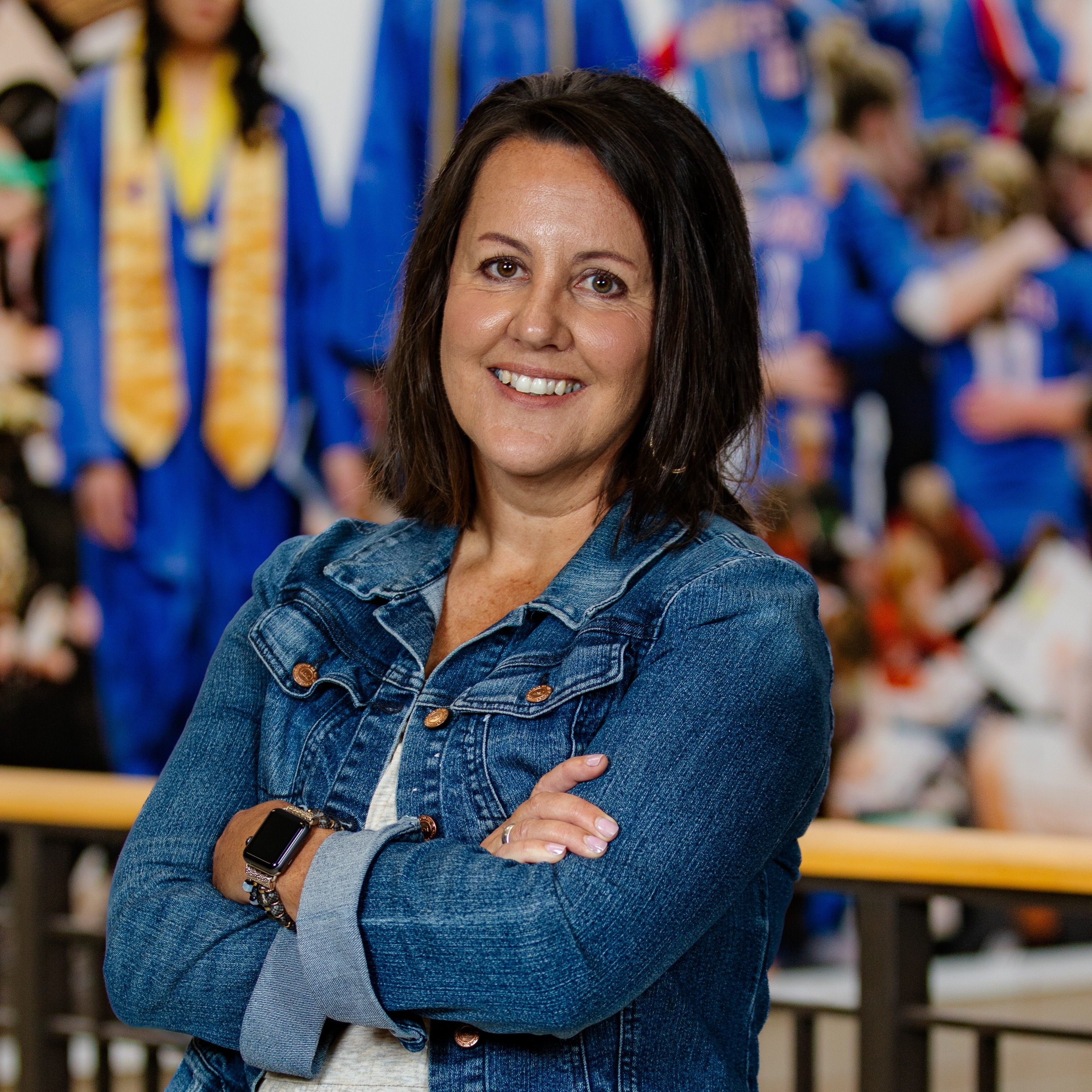 A smiling woman in a denim jacket with arms crossed, standing in front of a blurred graduation crowd.