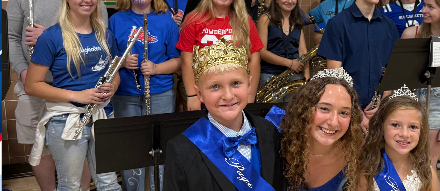 Homecoming court royalty and pages smile with high school band students holding instruments at a school event.