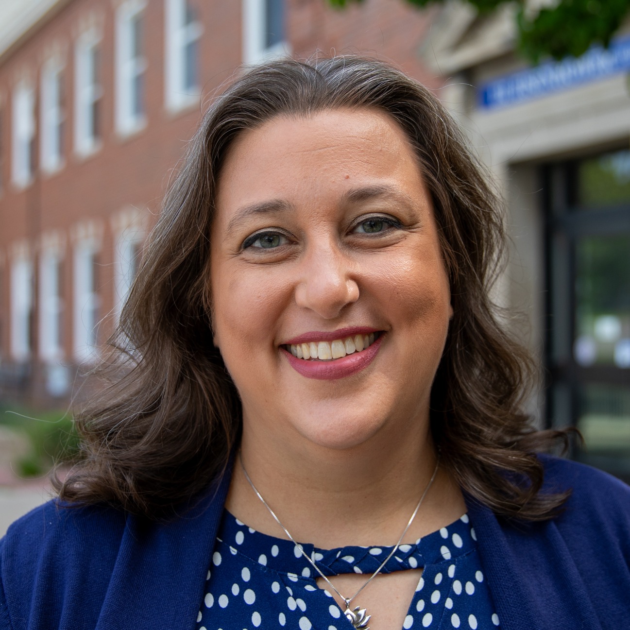 Debra Luna smiles for a photo. She is in a blue shirt and standing outside in front of a brick building.