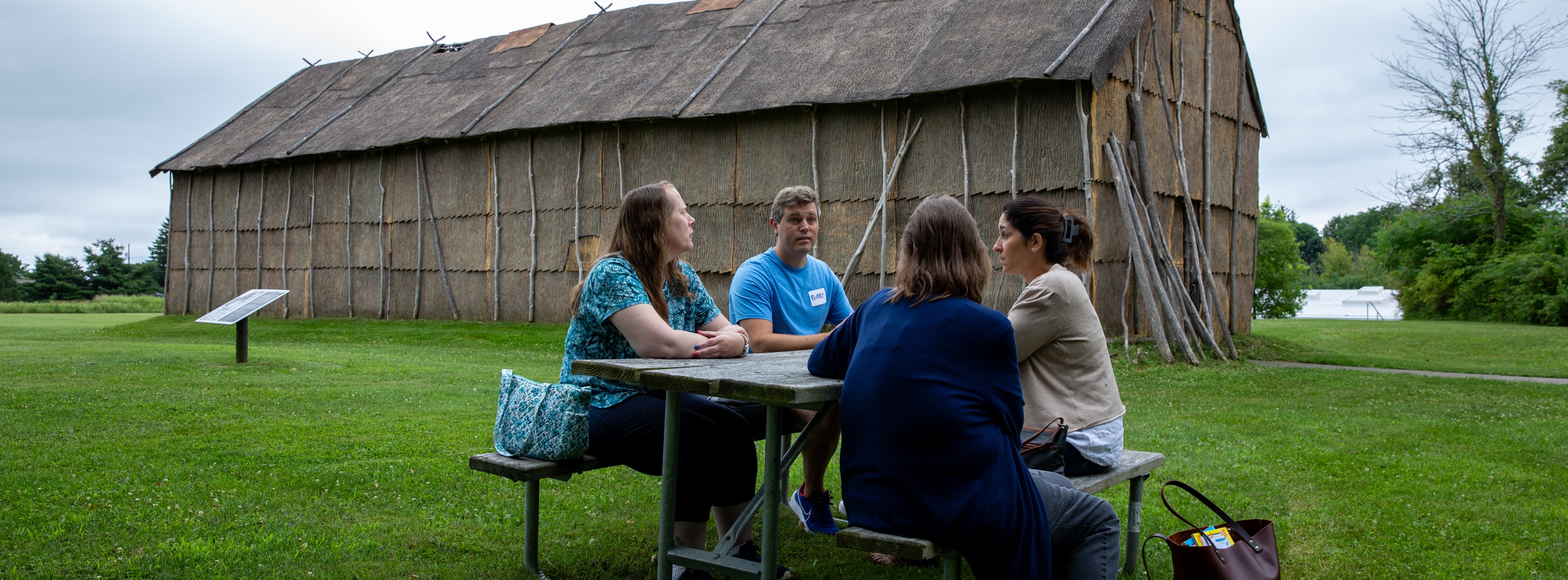 Four EduTech staff members sit around a picnic table near a longhouse at Ganondagan State Historic Site