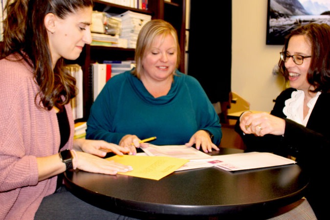 Three women sit around a table, reviewing documents. One points, another writes, and they all smile.