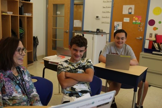 Three people in a classroom: two seated and one standing, all focused on a laptop. Desks, chairs, cabinets, and bulletin board visible.