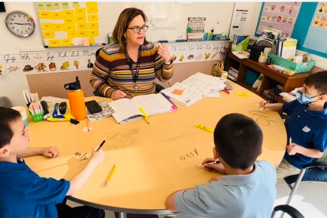 Teacher assists four young students around a table with papers, pencils, and markers in a classroom.