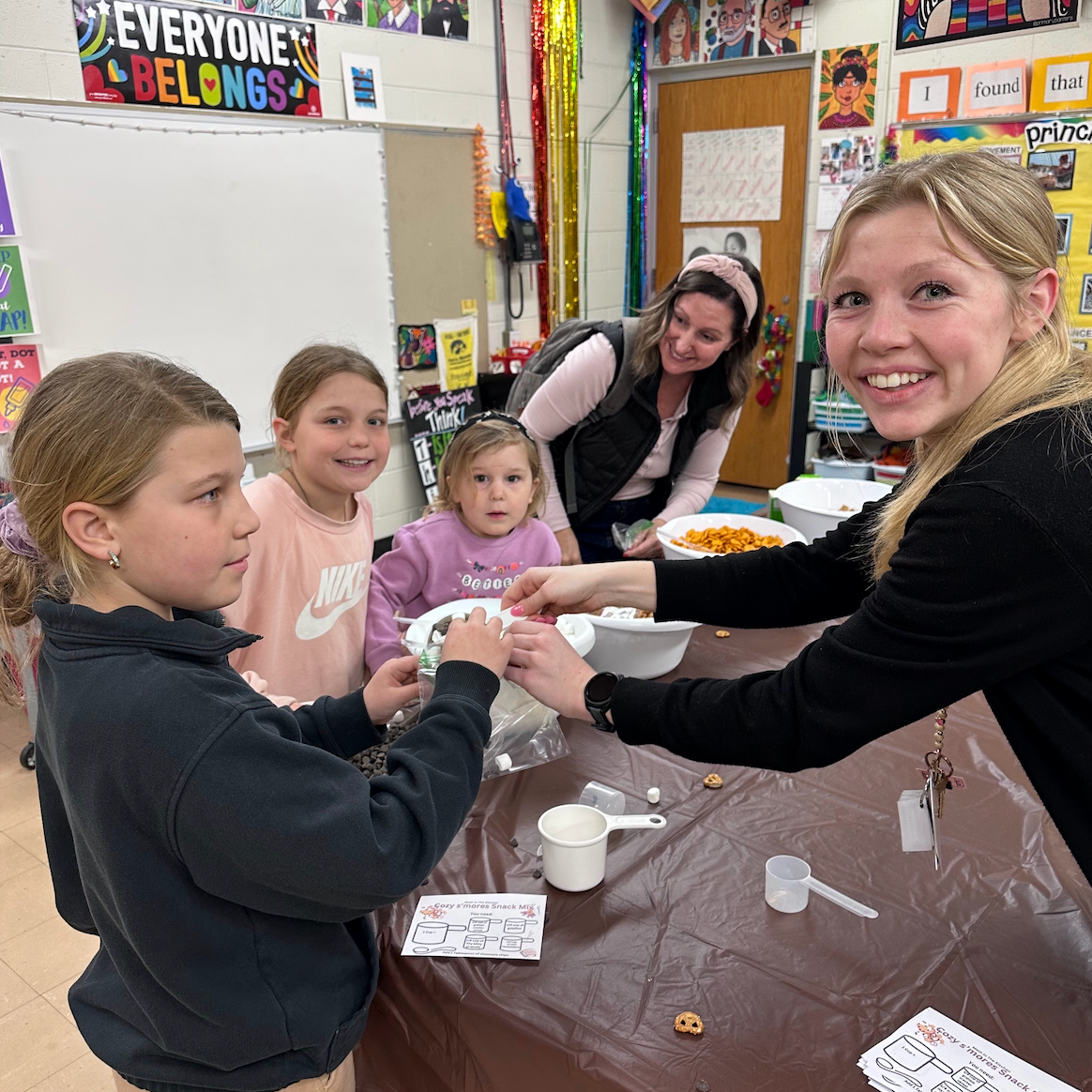 Two staff members serving dry ingredients into bowls for three female students for a chex mix snack