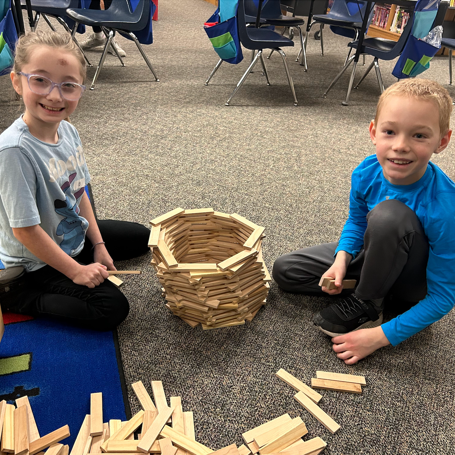 A female and male student sitting on the classroom floor building a circular tower with wooden planks