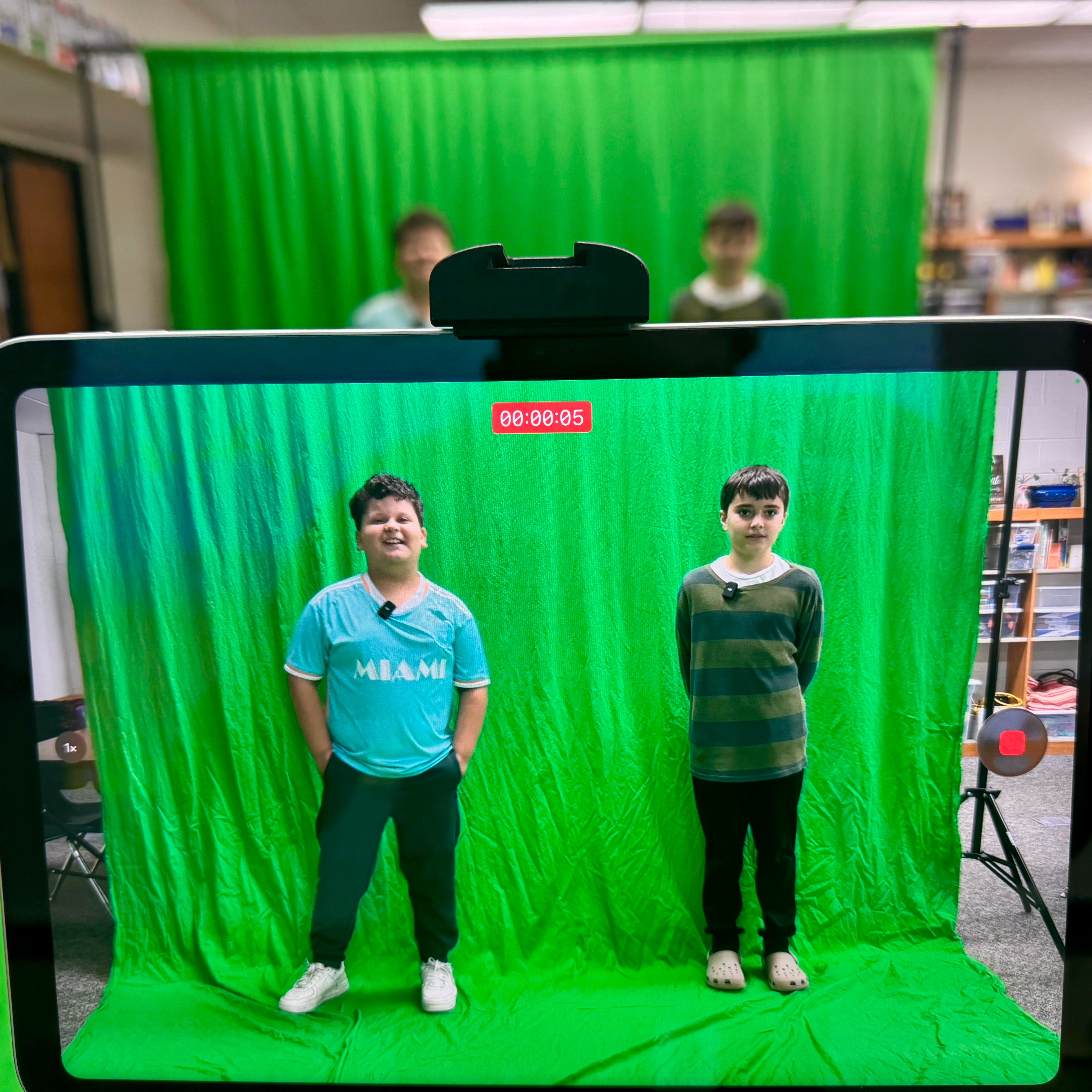 Two students standing in front of a green screen fabric wearing microphones being recorded through an ipad for a school news video