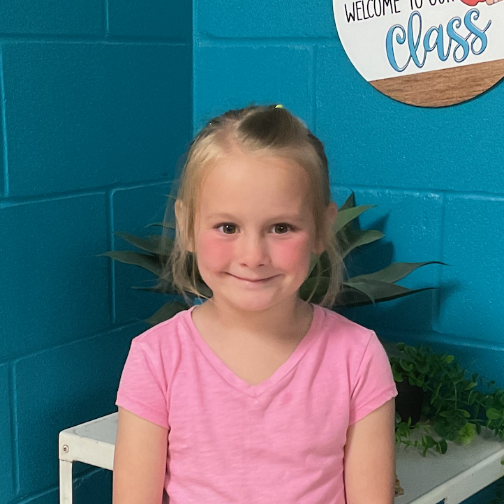 MP East student, Molly, standing in front of a turquoise wall with a sign that says Welcome to our class; wearing a pink shirt