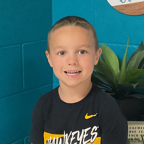 Miles Berg wearing a t-shirt that says HAWKEYES standing in front of a Welcome to our Class sign and a block that reads Teachers who love teaching, teach children to love learning