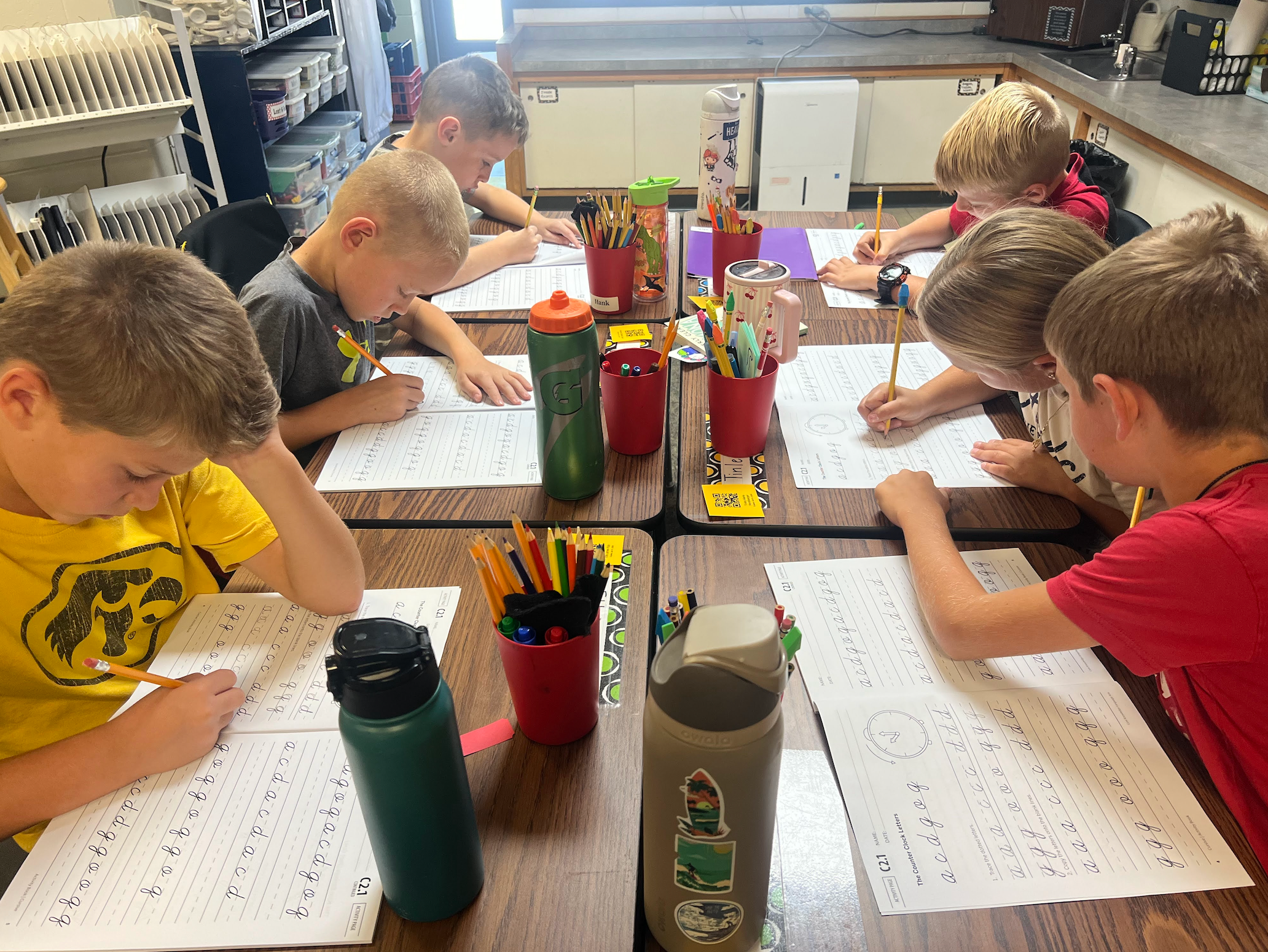 Students sitting at desks with pencils and a cursive practice sheet writing the letters a b c d e f g repeatedly