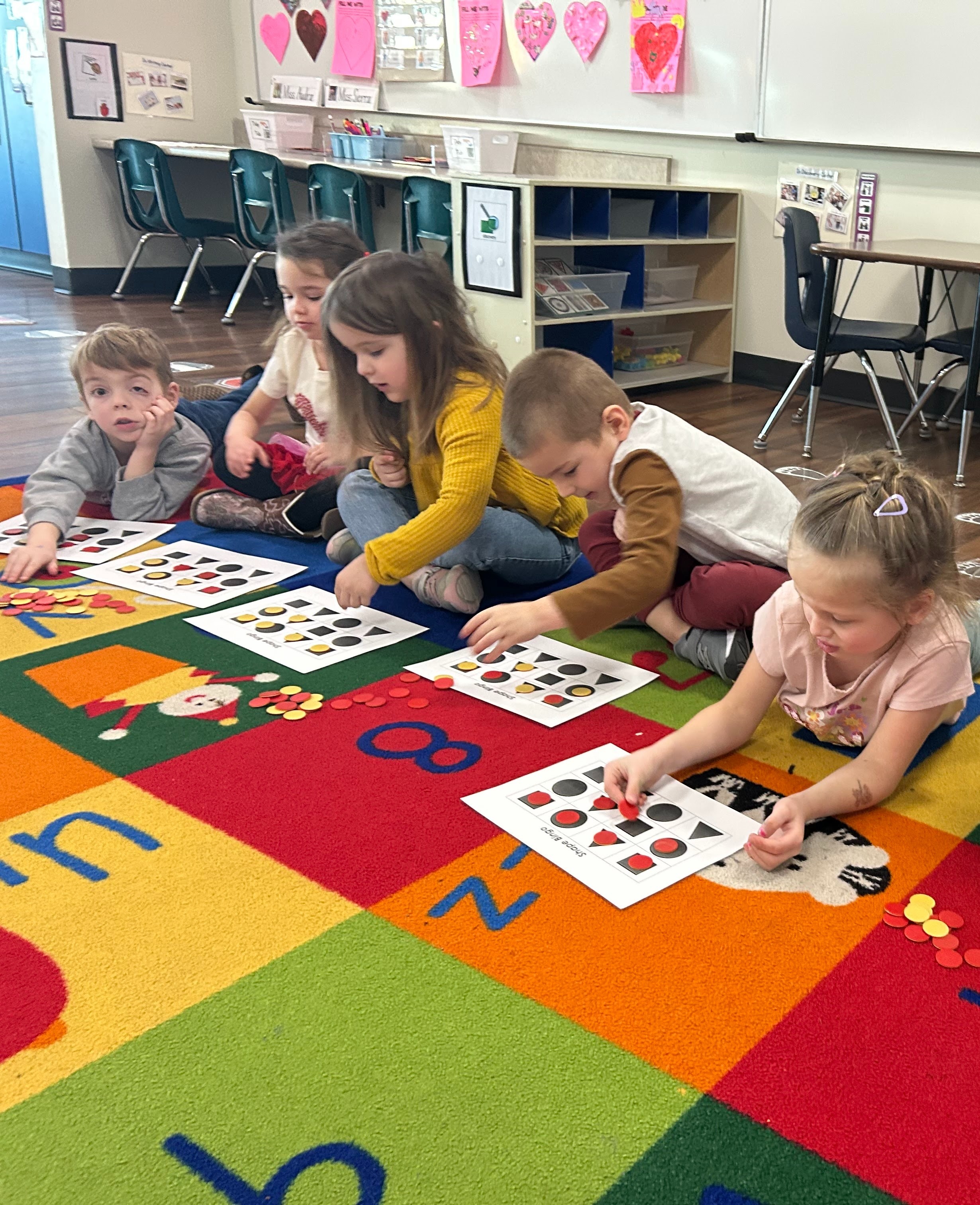 Preschool students playing shapes bingo sitting on the floor; bingo cards have shapes like square, triangle, circle, rectangle, students have red and yellow circular markers to cover each shape