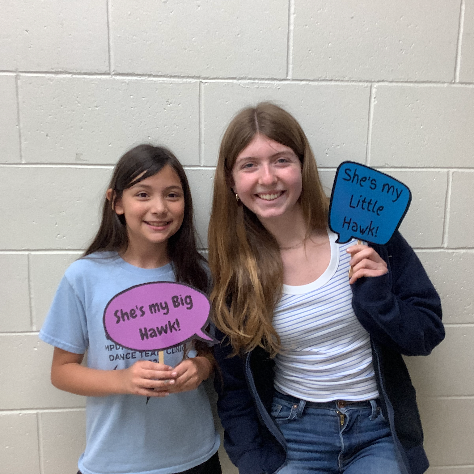 An elementary female student and a high school female student smiling together. They are holding signs reading: "She's my Big Hawk!" and "She's my Little Hawk!"