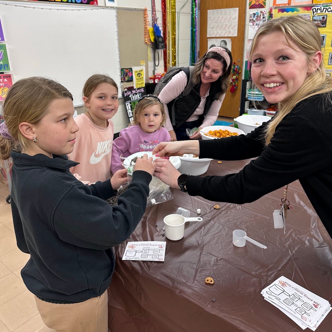 Two staff members dishing out dry ingredients into bowls for three female students for a chex mix snack