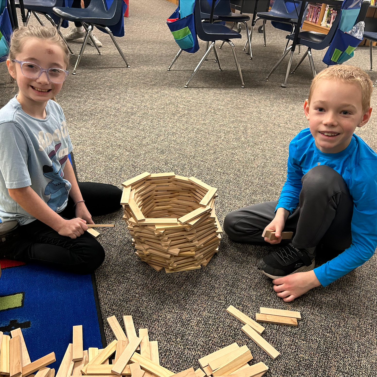 A female and male student sitting on the classroom floor building a circular tower with wooden planks