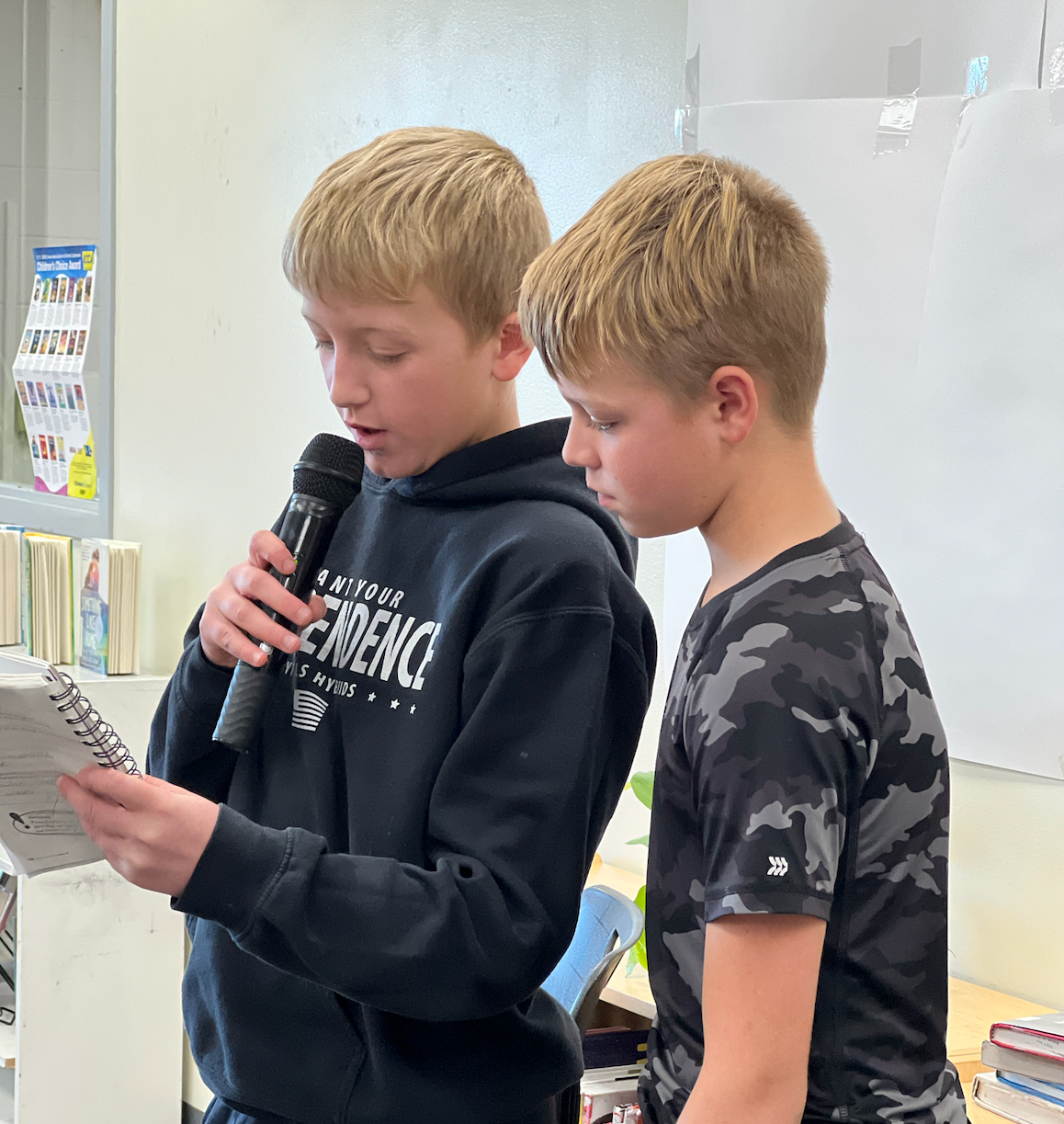 Two male students - one speaking into a microphone holding a notebook and the other looking over his shoulder at the notebook