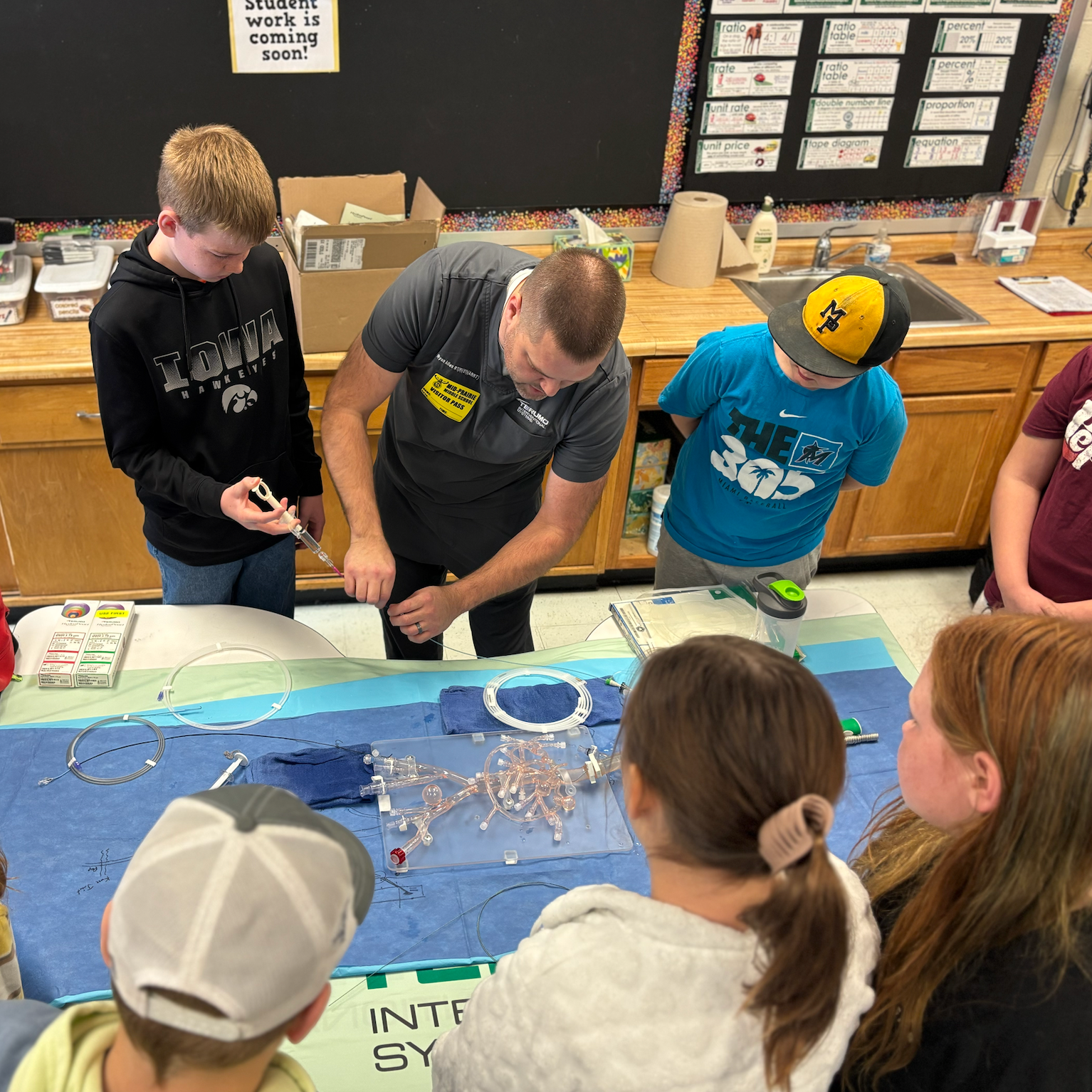 Volunteer demonstrating a science experiment for students; Students are watching, one student is holding a syringe for the experiment. Text on tshirts reads IOWA Hawkeyes and The 305