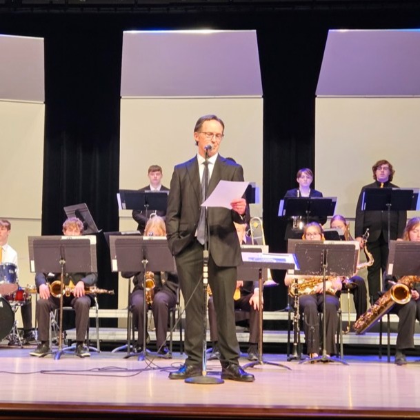 high school band members in chairs with music stands in front of them and band director David Kunz up front reading from a paper to the audience