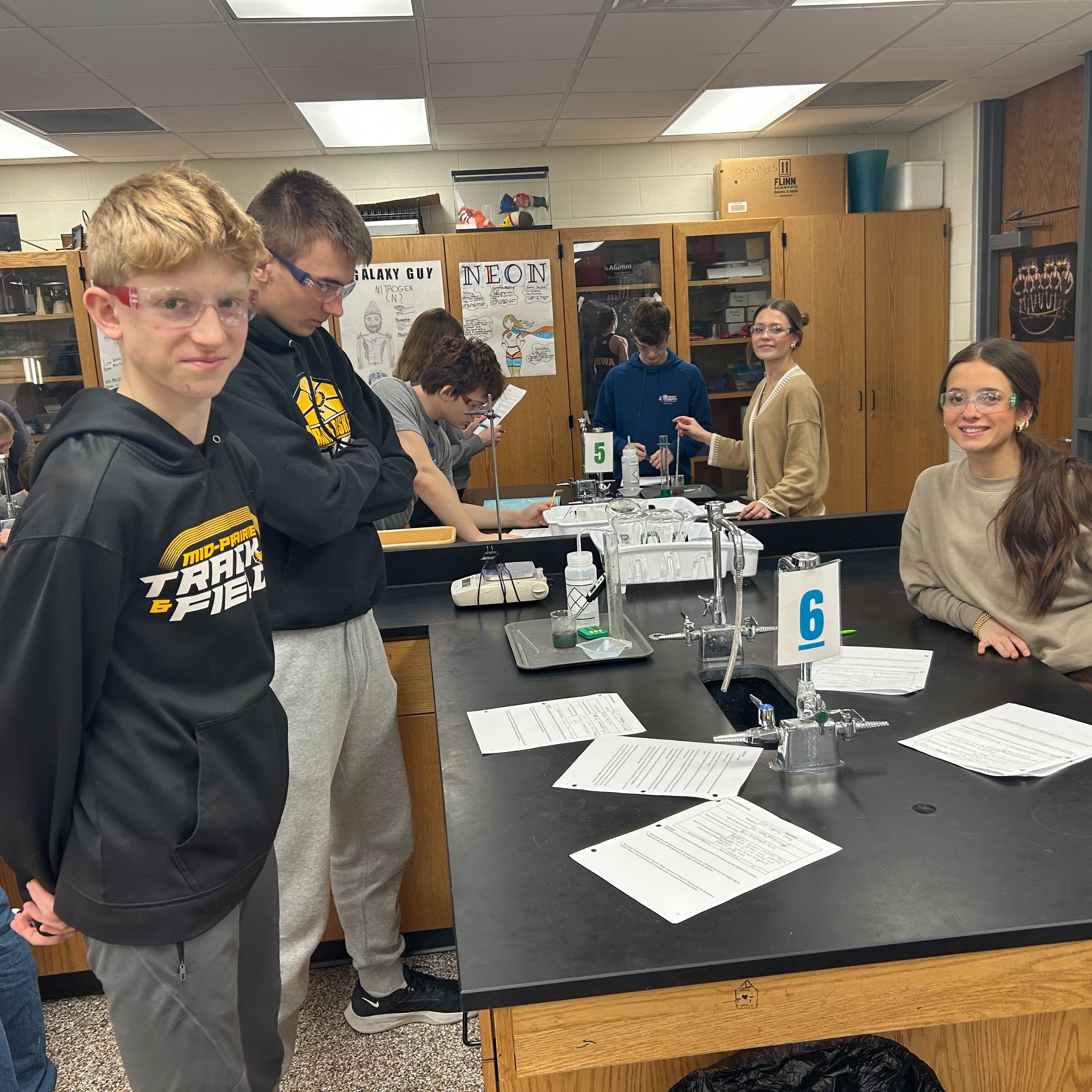 Students in a science lab with papers on the table smiling with science goggles on and the tables numbered 5 and 6