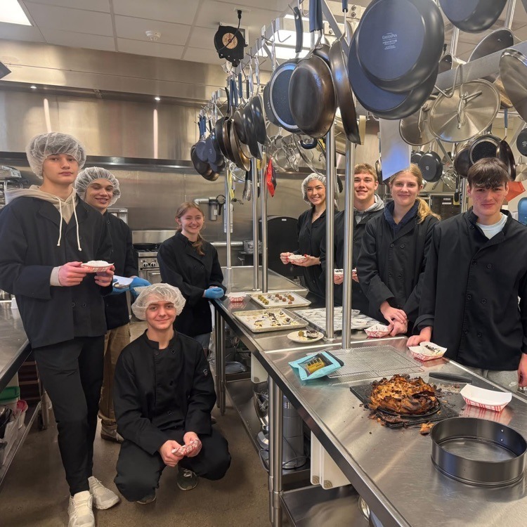 Students in the culinary lab with pots hanging overhead and plates of food on the steel tables wearing their black uniforms