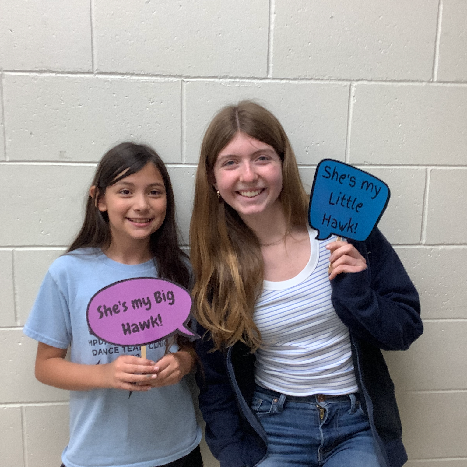 An elementary female student and a high school female student smiling together. They are holding signs reading: "She's my Big Hawk!" and "She's my Little Hawk!"