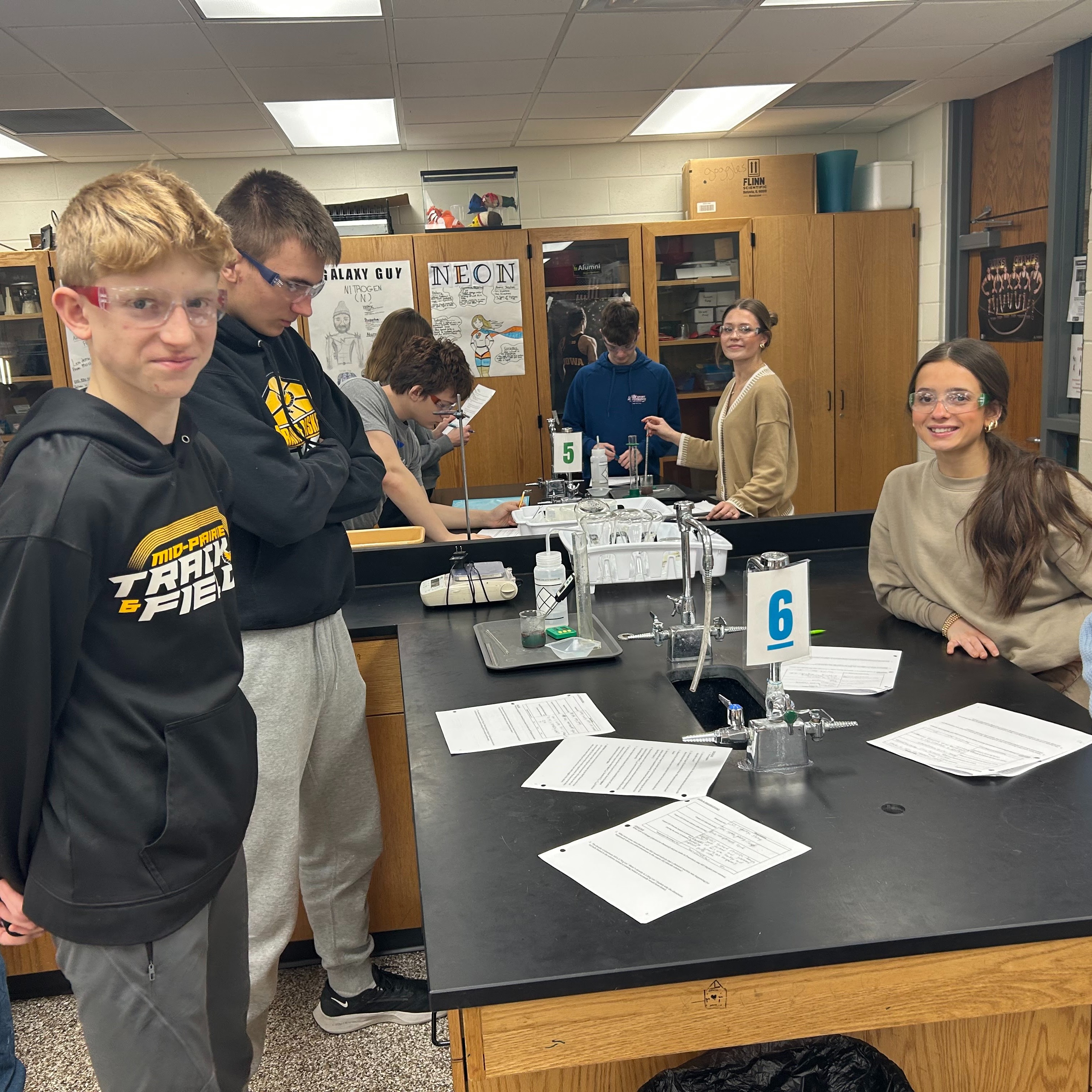 Students in a science lab with papers on the table smiling with science goggles on and the tables numbered 5 and 6