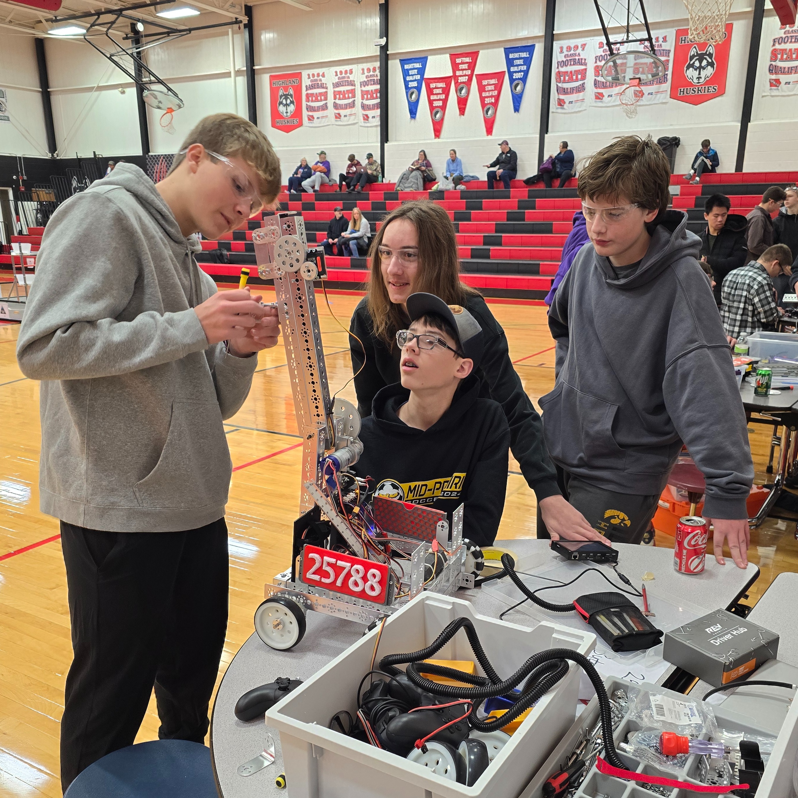 Four male students working on a robot they built; Three students are watching while one student attaches something to the arm of the robot that reaches up; Robot has the numbers 25788 on it
