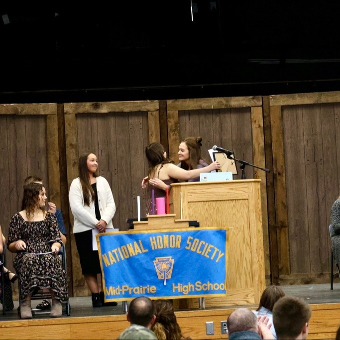 Students seated to the left and staff members seated to the right; One student giving a staff member a hug up at the podium on stage with a sign in front that reads National Honor Society Mid-Prairie High School