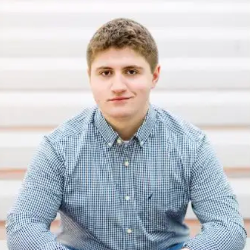 Miles Edgington  senior photo sitting outdoors in front of white steps wearing a blue plaid button up shirt