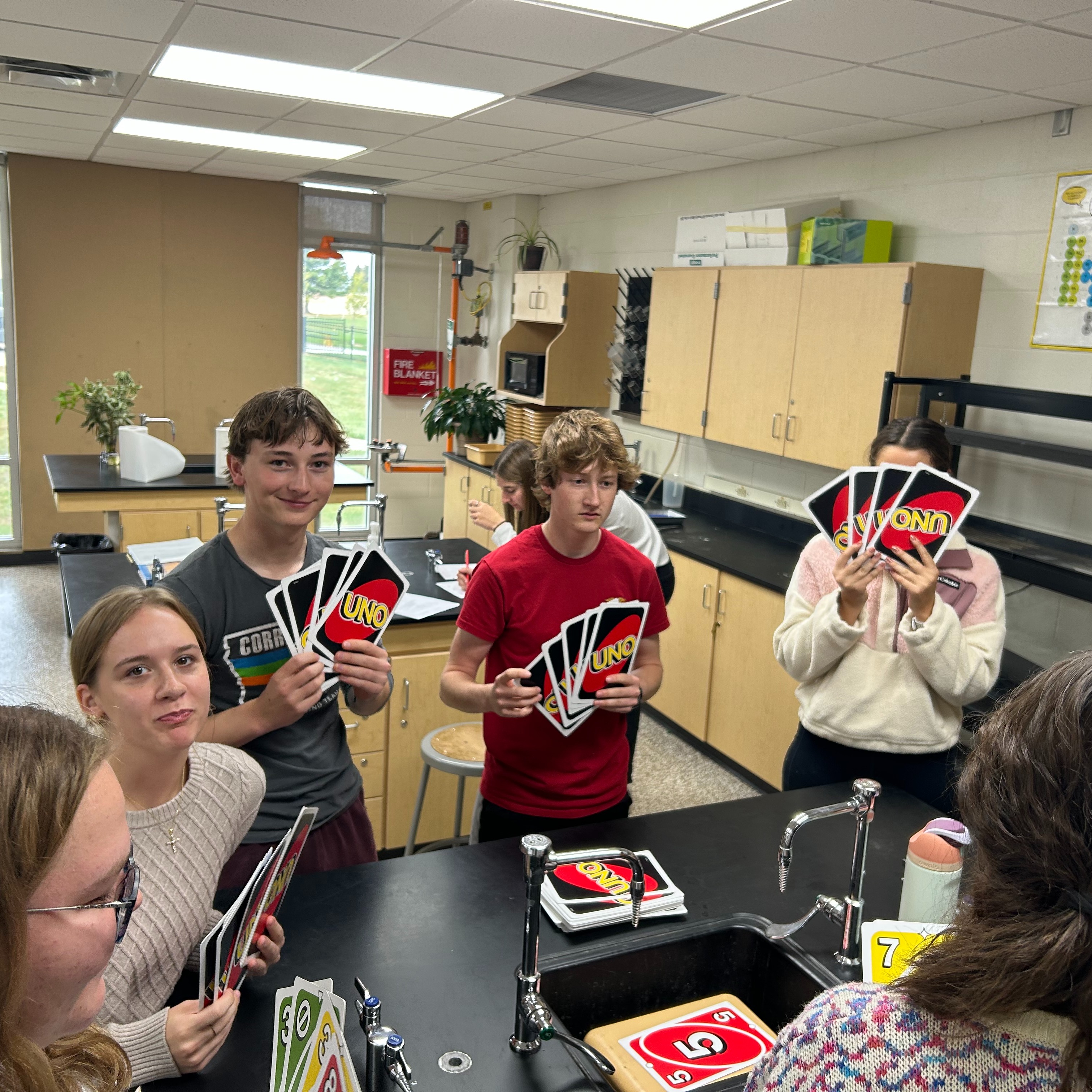 Group of 7 high school students playing UNO with large-sized uno cards, two are smiling, one is looking at the camera, one is covering her face with the cards, and others are looking at the stack int he center of the table.