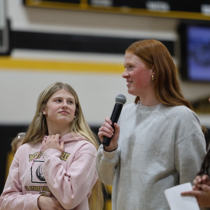 Two female students standing in the high school gym, one is holding a microphone and speaking into it smiling, the other is smiling looking at the girl with the microphone