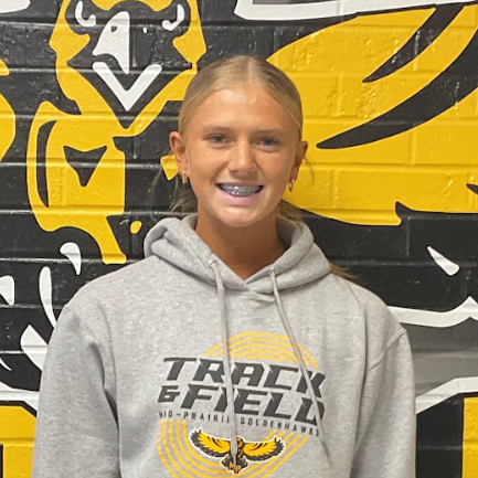 Tessa wearing a grey sweatshirt that says Track and Field standing in front of the black and gold MP hawk wall decal