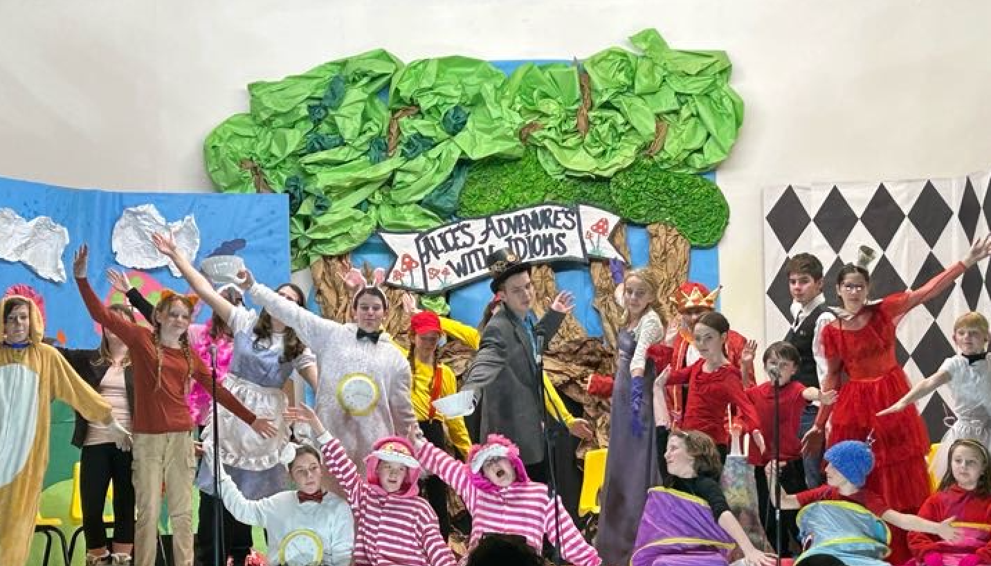 Students on stage with theater set background for Alice in Wonderland and in costume with arms up singing a song; text reads Alice's Adventures with Idioms
