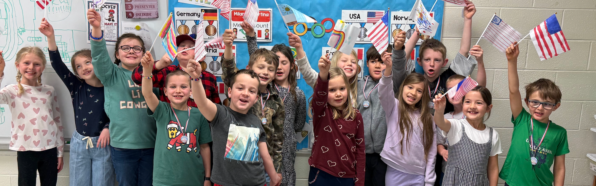 Students standing as a group in the classroom smiling all holding up a miniature American flag; text on wall behind them reads Milan Italy Let the Games Begin 2026