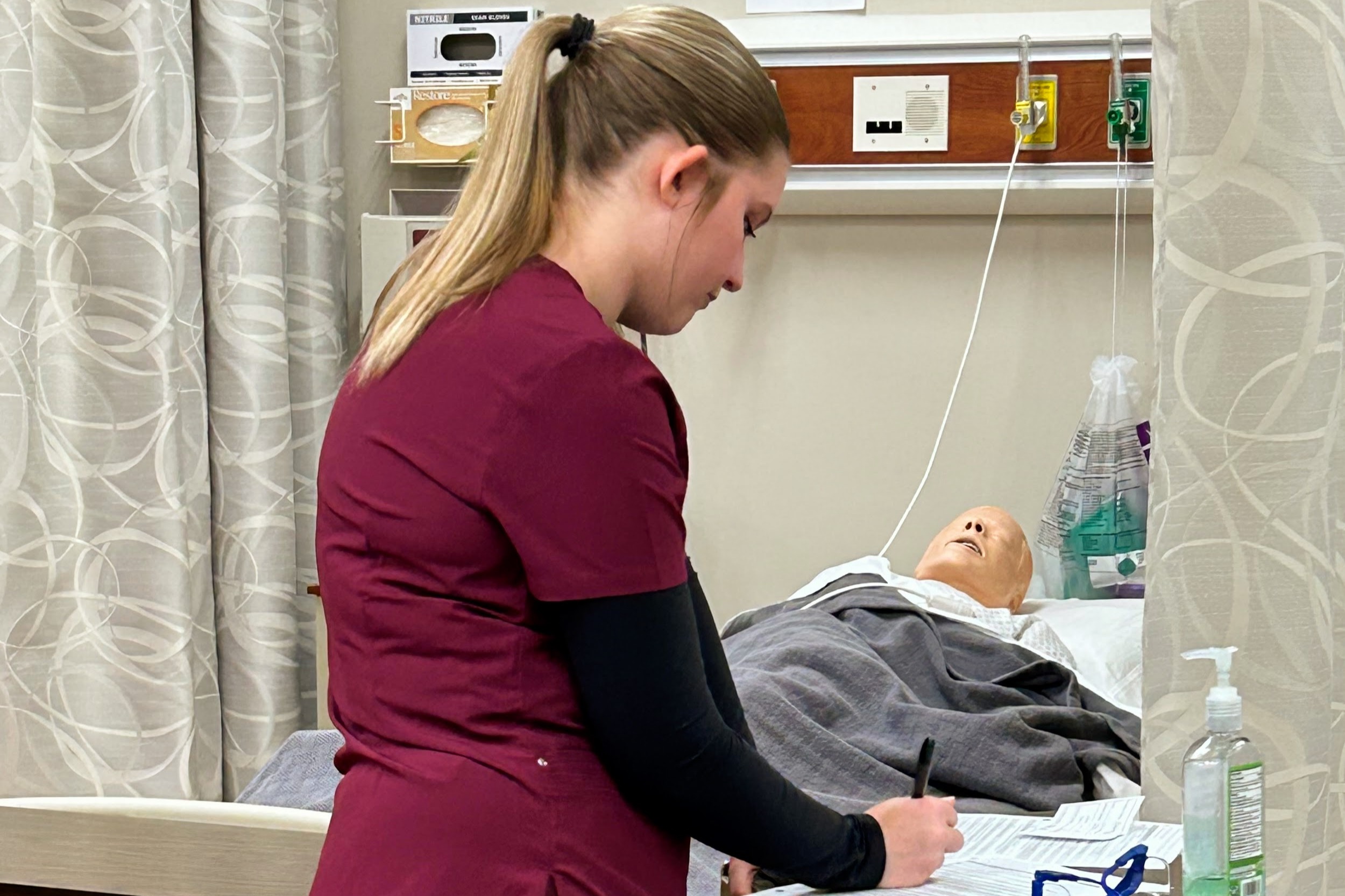 Nursing student writing on medical chart, with patient in the background.