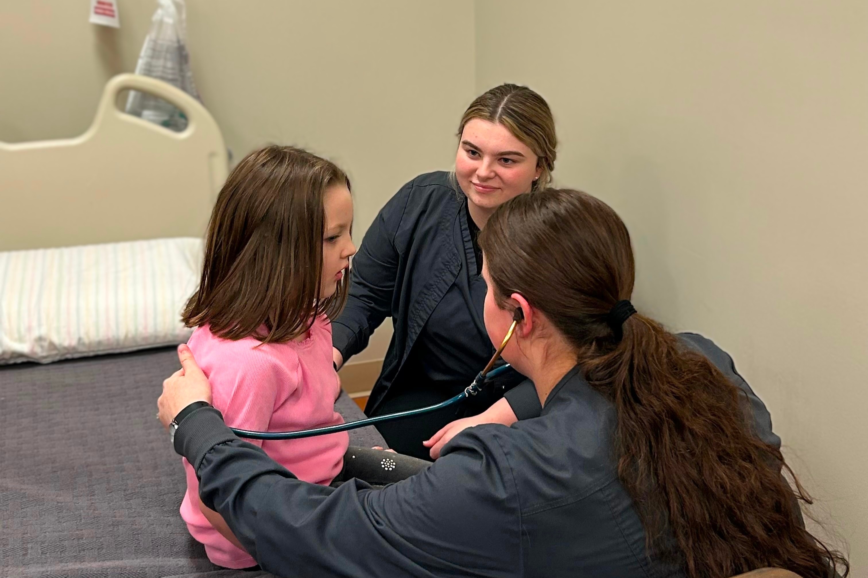 Nursing student with stethoscope listening to a young girls back. Another nursing student watching.