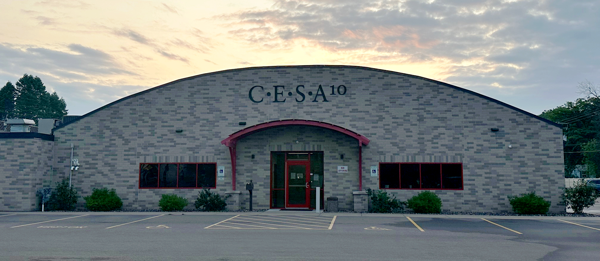 Exterior view of the C•E•S•A 10 building, a single-story grey brick structure with an arched roofline and a red-framed entrance.