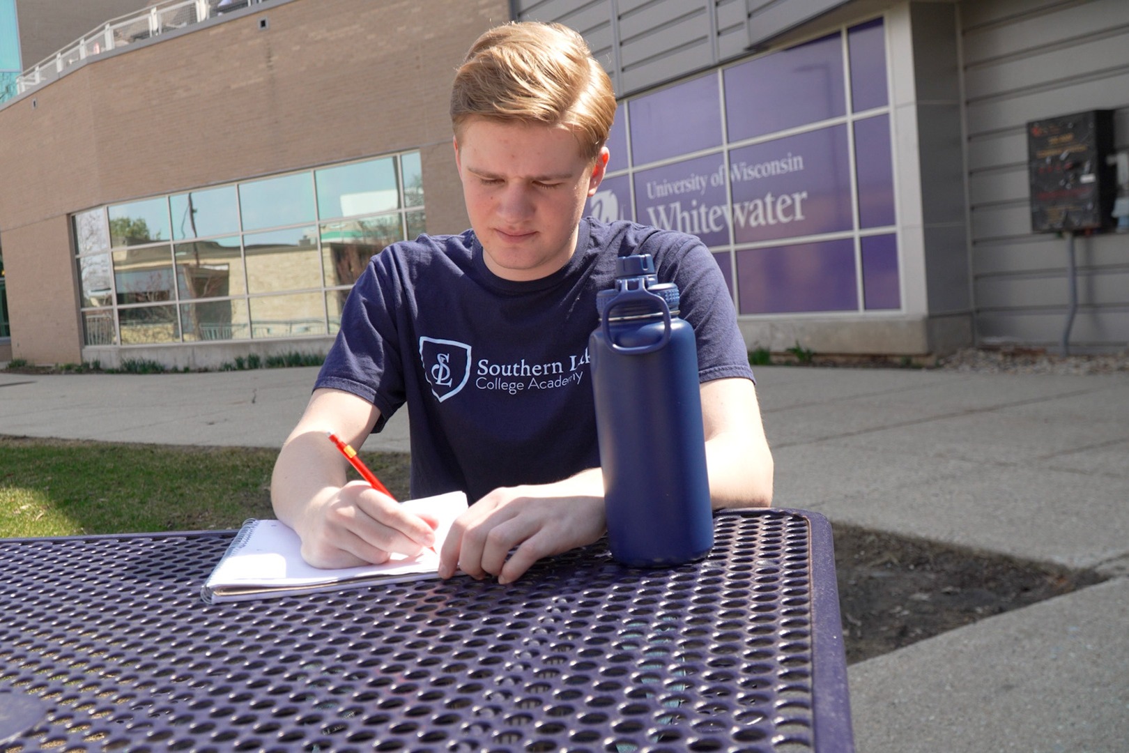 A man in a blue shirt writes on a piece of paper with a red pen at a table.