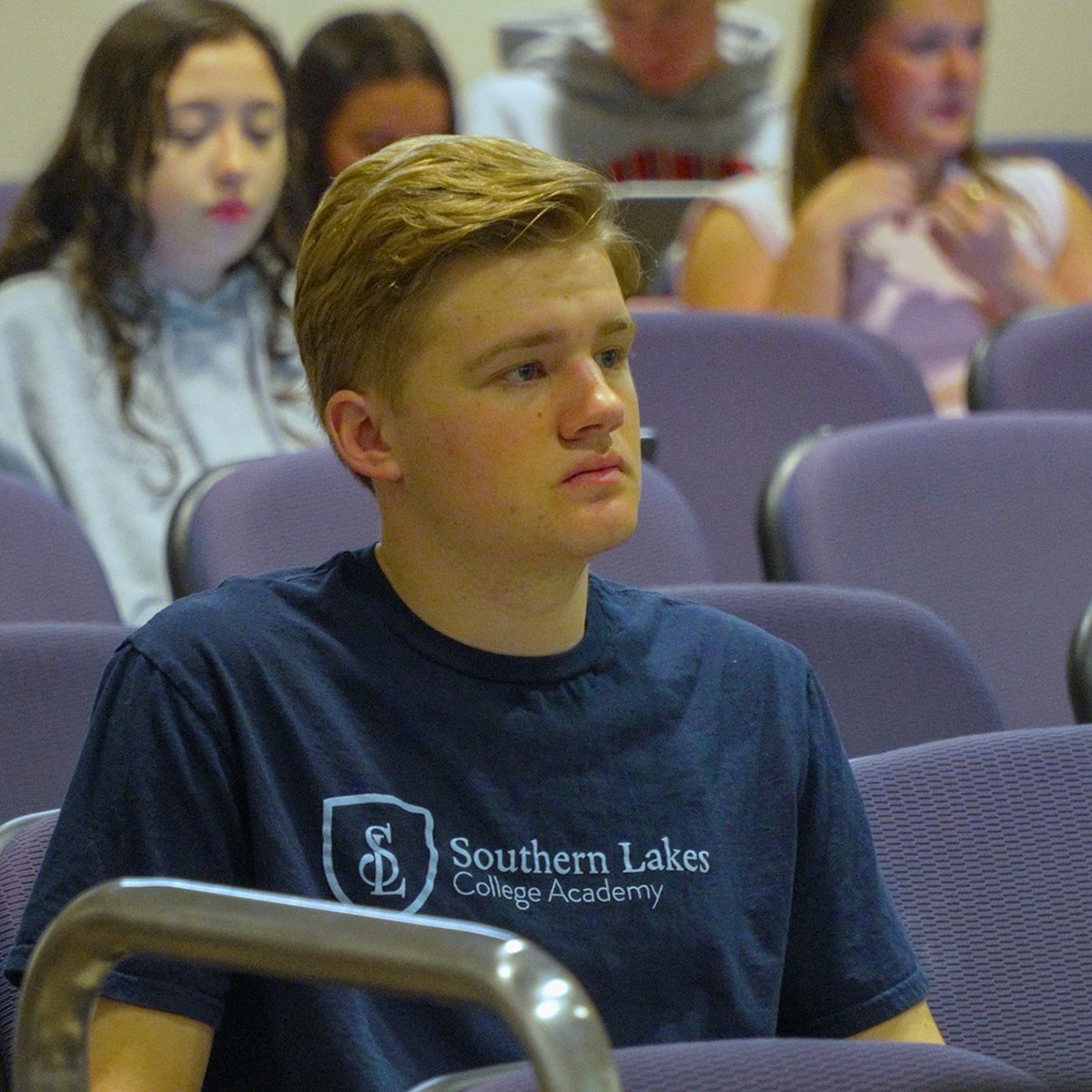 A student in a blue T-shirt sits in a lecture hall, with other students seated behind him.