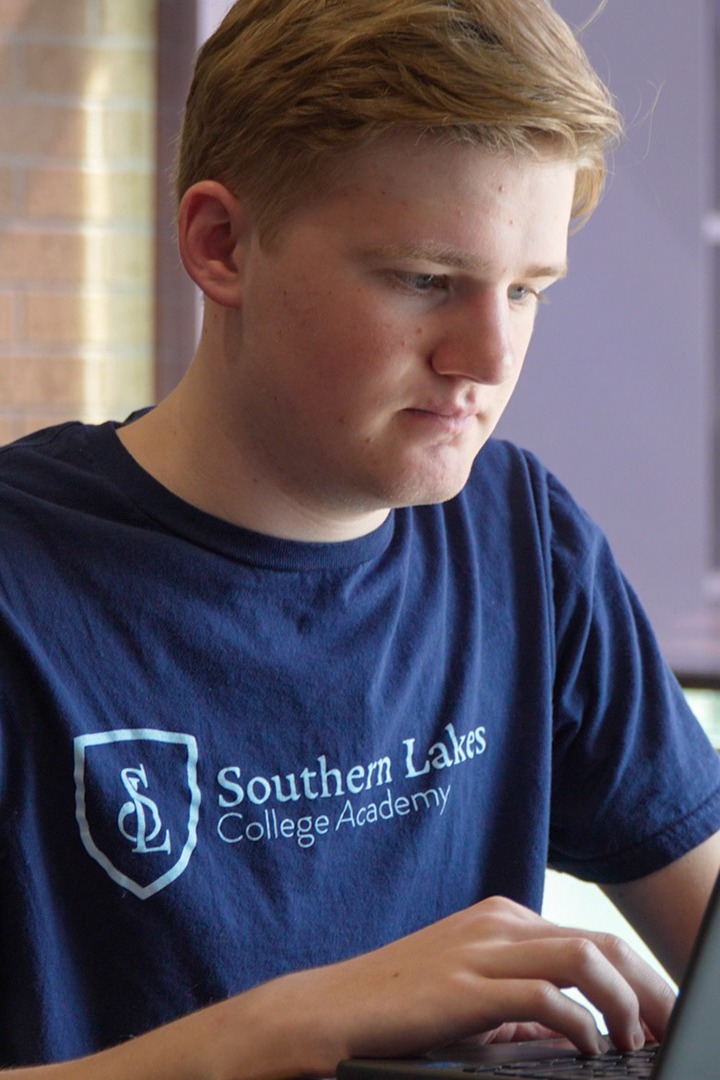 A young man, wearing a navy blue t-shirt with a logo, sits at a table with a laptop.
