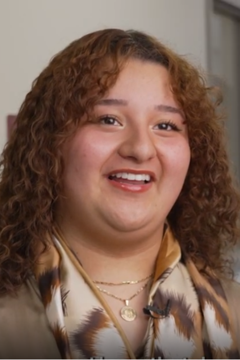 A woman with curly hair smiles, wearing a scarf and a necklace. She is standing in a hallway with a window in the background.