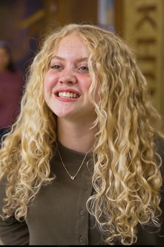 A woman with curly blonde hair smiles at the camera, wearing a brown top and a necklace.
