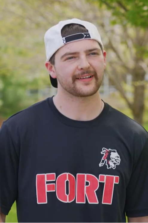 A man in a white hat and black T-shirt with "FORT" in red letters stands on a lawn.