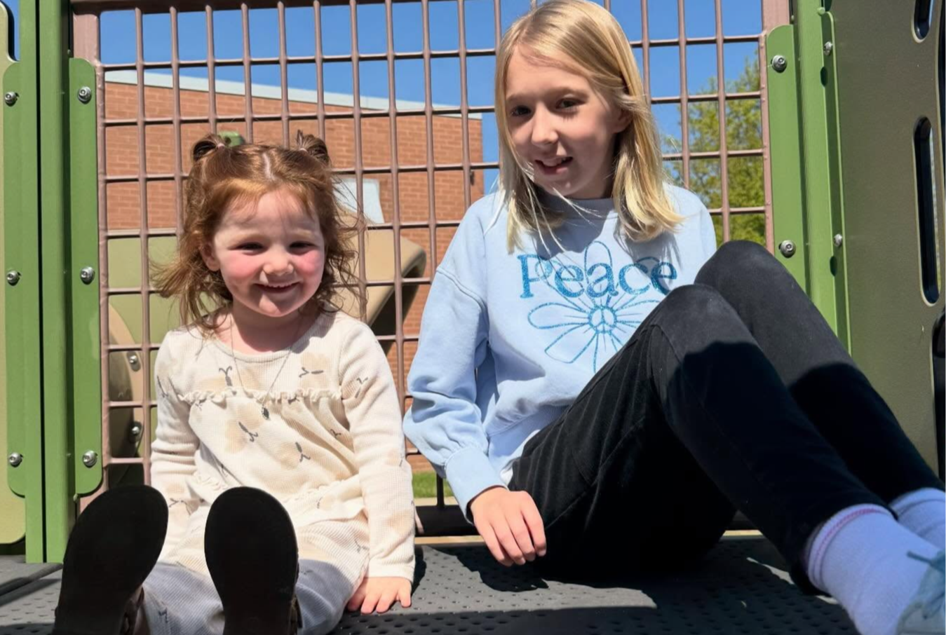 Two young girls sit outdoors in a playground, one smiling. They sit near a green gate.