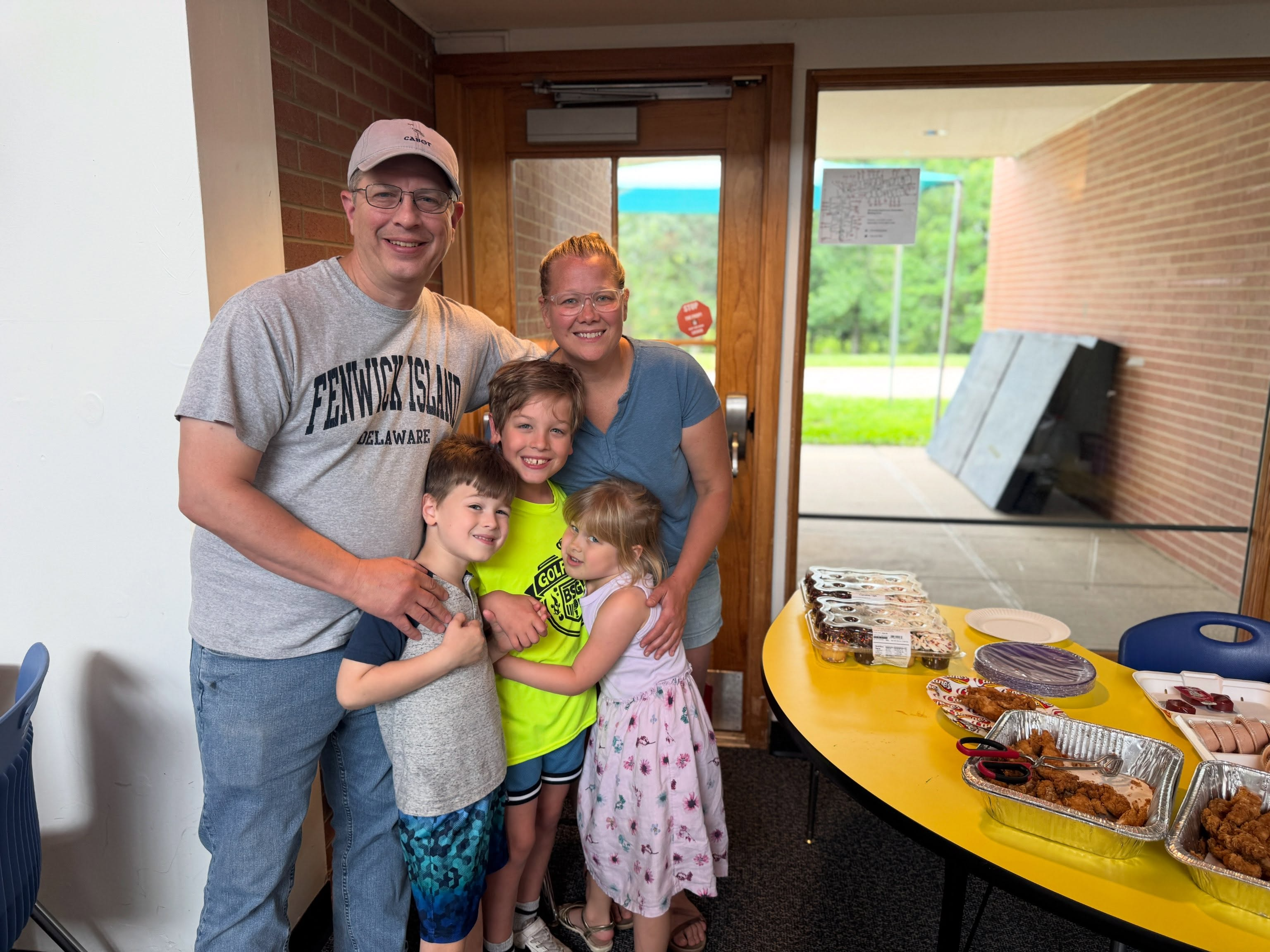 Family stands together in a room with a yellow table and food. Three children are smiling.