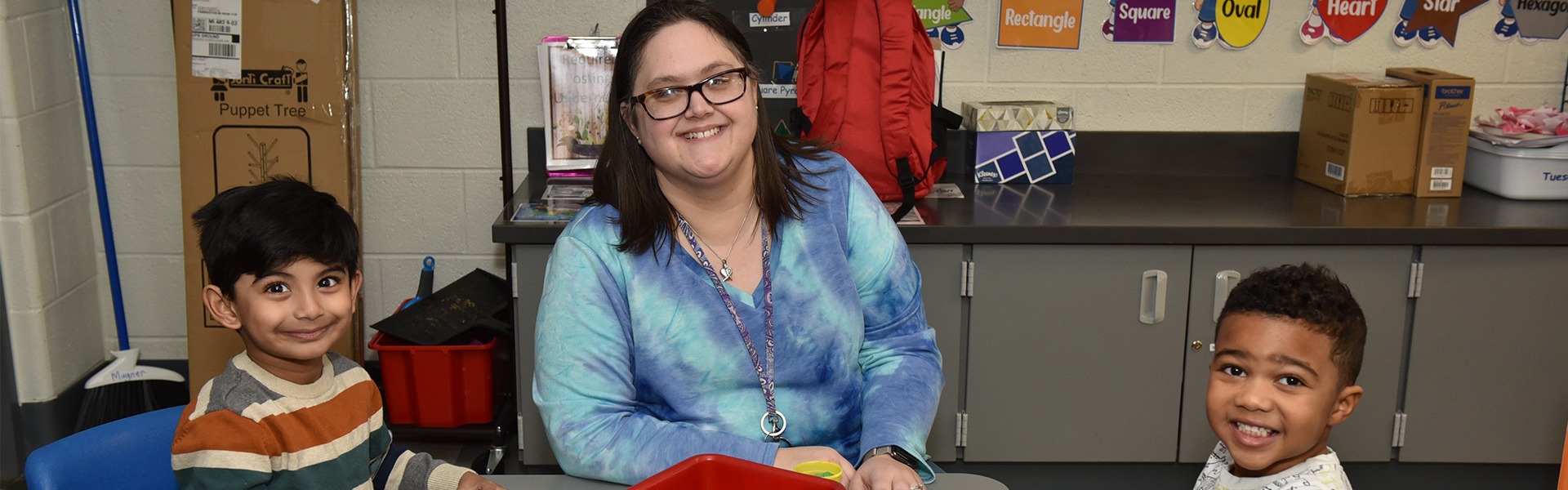 Teacher and two children sitting at a table with red bins filled with toys in a classroom setting.