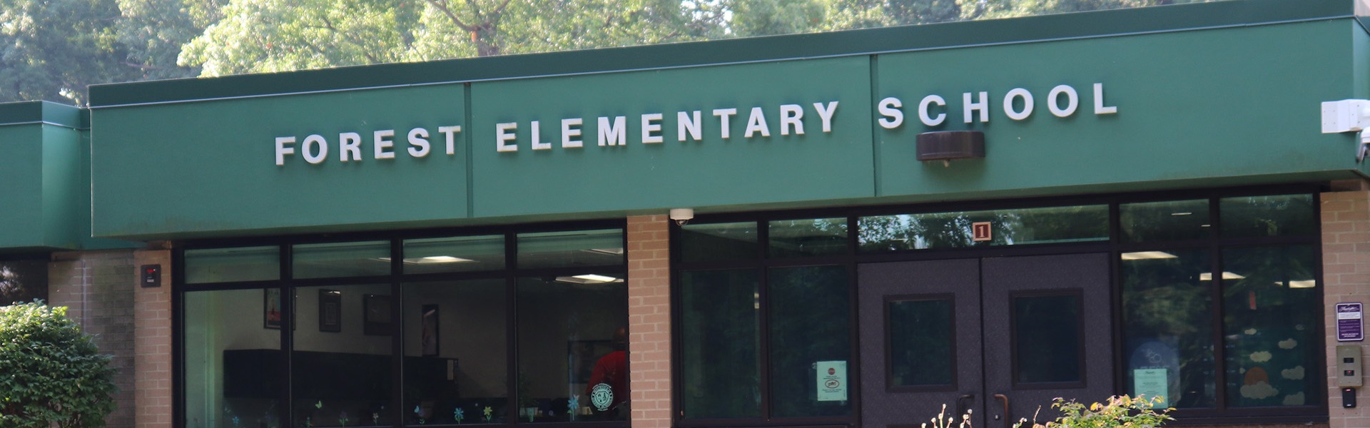 Front entrance of Forest Elementary School with green signage above large windows and a bench in the landscaped garden area.
